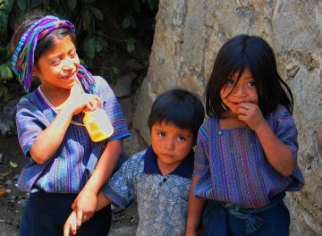 Three Guatemalan children in the colorfully-woven clothing of the native peoples shyly smile at the camera.